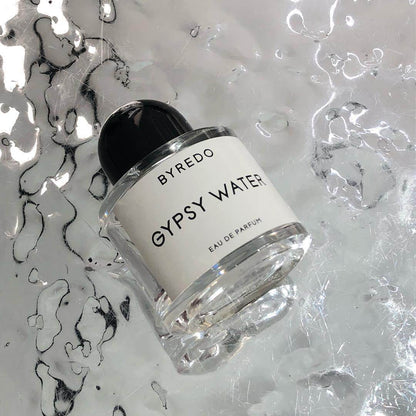 A bottle of Baccarats Gypsy Water EDP For Men and Women. The transparent cylindrical glass bottle features a white label with black text and a black rounded cap. Embracing the essence of nomadism, the fragrance name "Gypsy Water" and "Eau de Parfum" are elegantly displayed on the label.