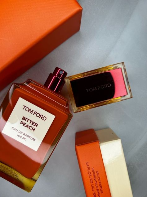 A 100 ml bottle of Baccarats Tom Ford Bitter Peach Eau De Parfum for Men and Women features a warm orange hue that reflects the luxuriant notes of Sicilian blood orange oil and sandalwood absolute, with a square label displaying both the brand and fragrance name.