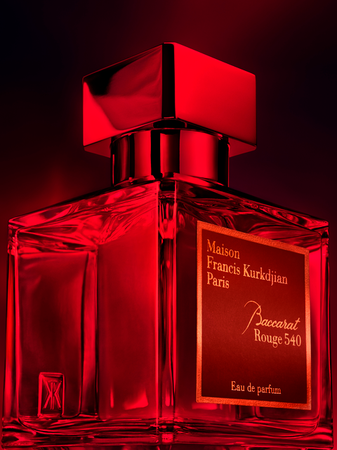 A transparent glass bottle of perfume with a gold cap, featuring "Baccarats Baccarat Rouge 540 Eau de Parfum for Men and Women" in gold and white text on a red background, embodies the essence of an amber floral fragrance.