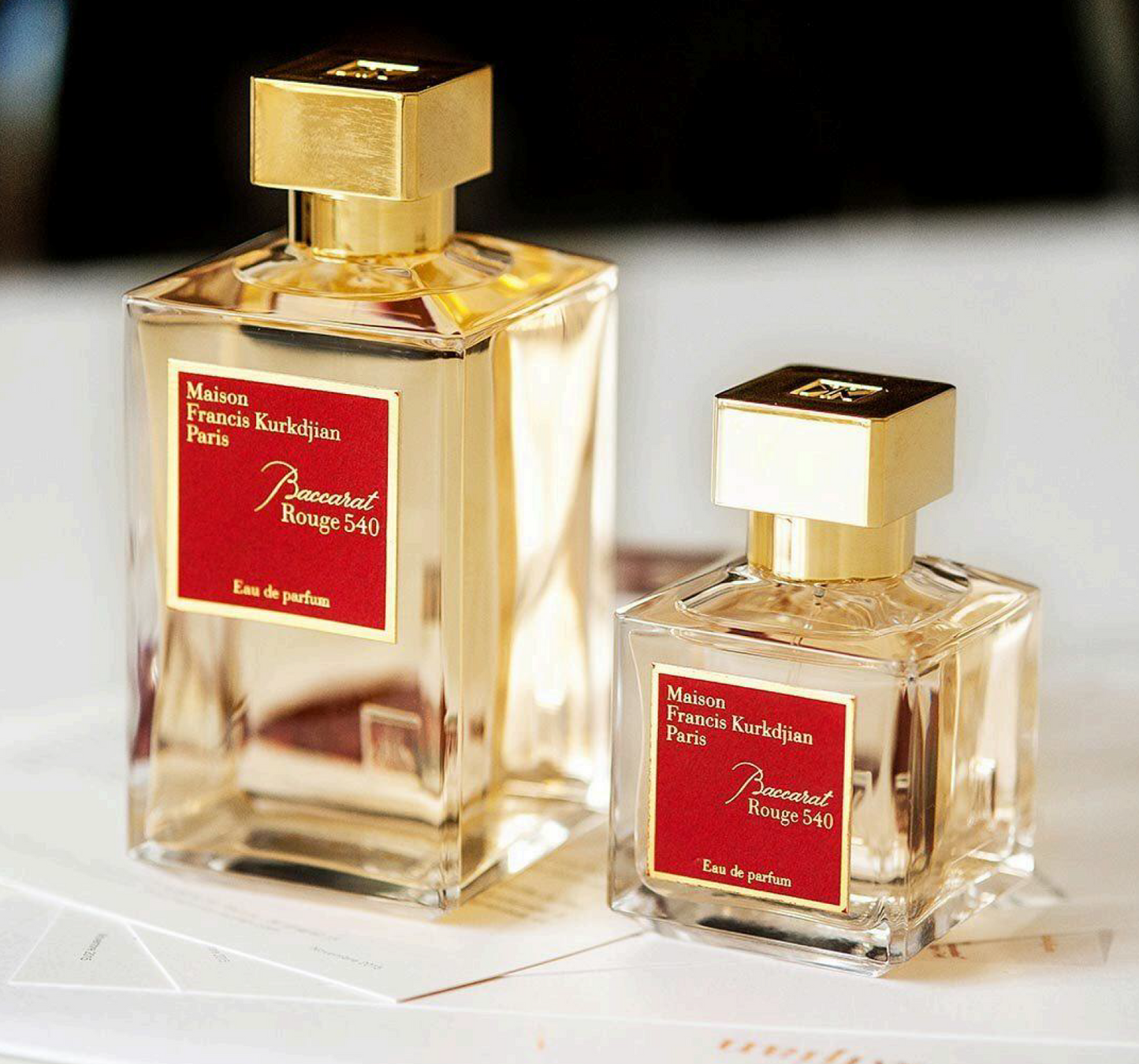 A transparent glass bottle of perfume with a gold cap, featuring "Baccarats Baccarat Rouge 540 Eau de Parfum for Men and Women" in gold and white text on a red background, embodies the essence of an amber floral fragrance.