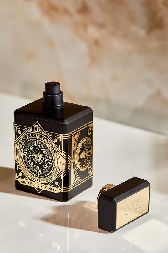 A 90ml bottle of Baccarats' Initio Oud for Greatness EDP. The black bottle, adorned with elaborate gold detailing, features an intricate design highlighted by a prominent eye motif on the front. Infused with rich and captivating notes of patchouli and oud wood, the fragrance exemplifies luxury. The cap is black with elegant gold accents.