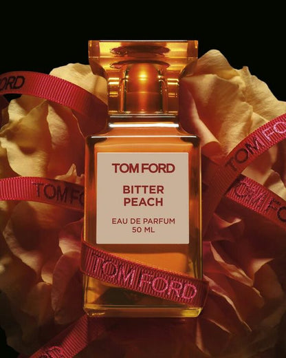 A 100 ml bottle of Baccarats Tom Ford Bitter Peach Eau De Parfum for Men and Women features a warm orange hue that reflects the luxuriant notes of Sicilian blood orange oil and sandalwood absolute, with a square label displaying both the brand and fragrance name.