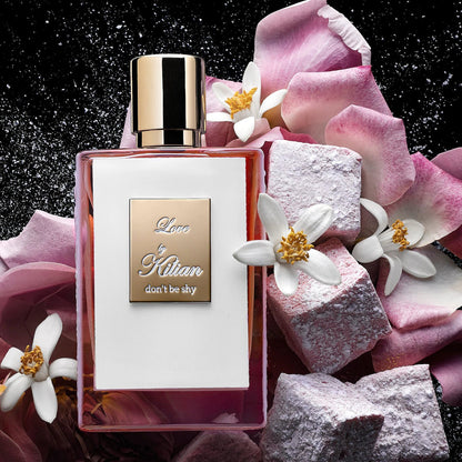 A rectangular glass bottle of By Kilian Love, Don't Be Shy perfume with a gradient pink hue, featuring a gold cap and a square gold label on the front, embossed with the text "Love by Kilian, don't be shy." Infused with delicate notes of Orange Blossom, this Baccarats fragrance captivates from the first spritz.